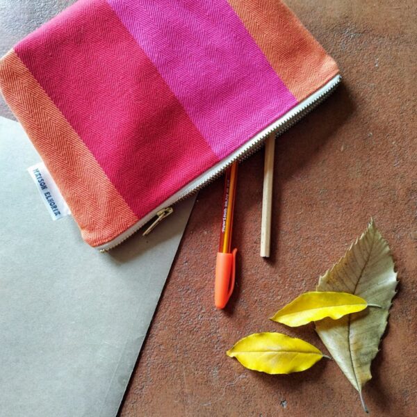 red orange pink srtriped bag flat on the floor with 2 pen coming out of it and a few leaves