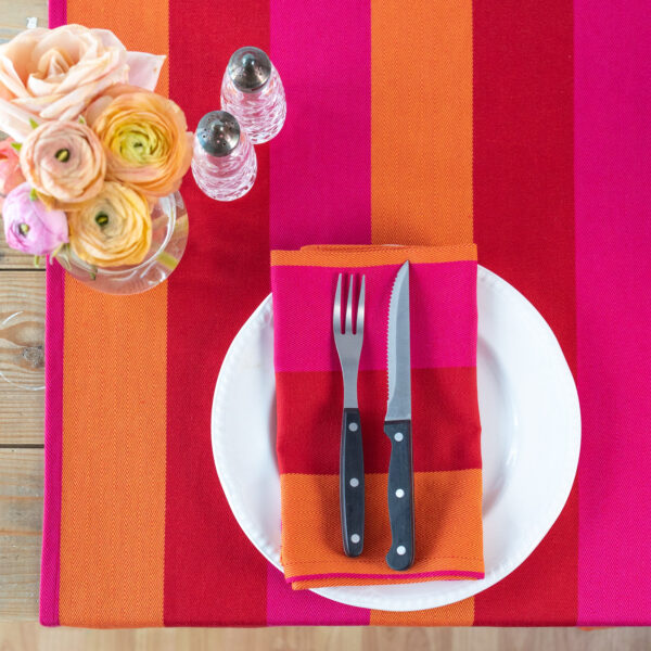 red orange pink napkin folded on a white plate with knife and fork on it, on a wooden table and red orange pink table runner with flowers and salt and pepper in the top left corner